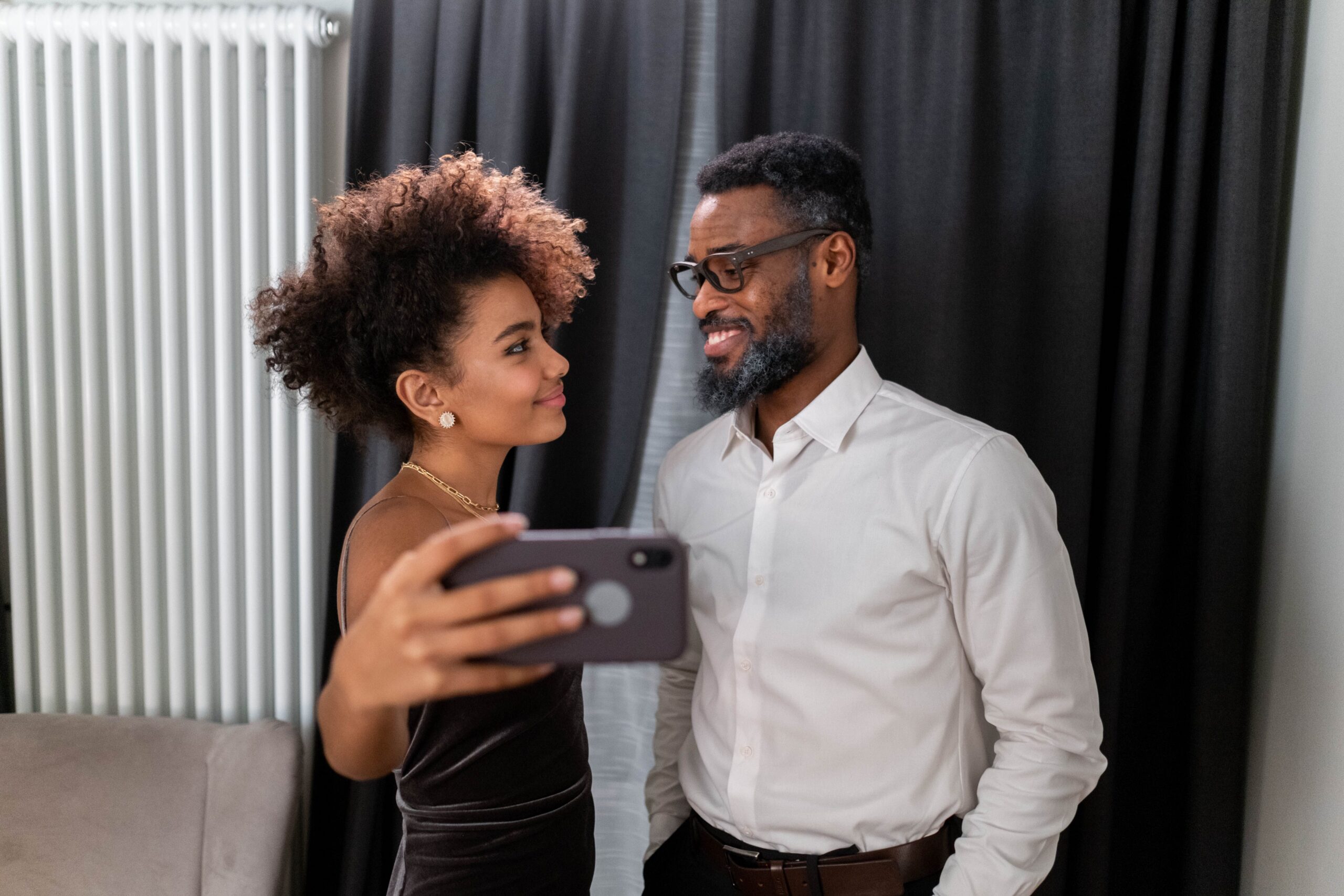 A happy father and daughter take a cheerful selfie indoors against a curtain backdrop.